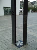 A memorial in one tile of a public square. The base is a black tile with five large circles around five small circles around a smaller circle. White stone shows through the circles. Out of opposite corners rise two very tall L-shaped columns. One column is pierced with the same circle pattern. The other column bears the inscription in the text.